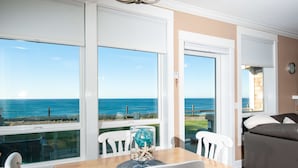 Un espace repas offrant une vue sur la mer, avec des chaises blanches, une table en bois et une table en verre surmontée d’objets décoratifs.
