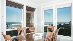 Un espace repas avec une table en verre et des chaises en bois, de grandes fenêtres offrant une vue sur l’océan, ainsi qu’une vue sur la plage et l’océan.