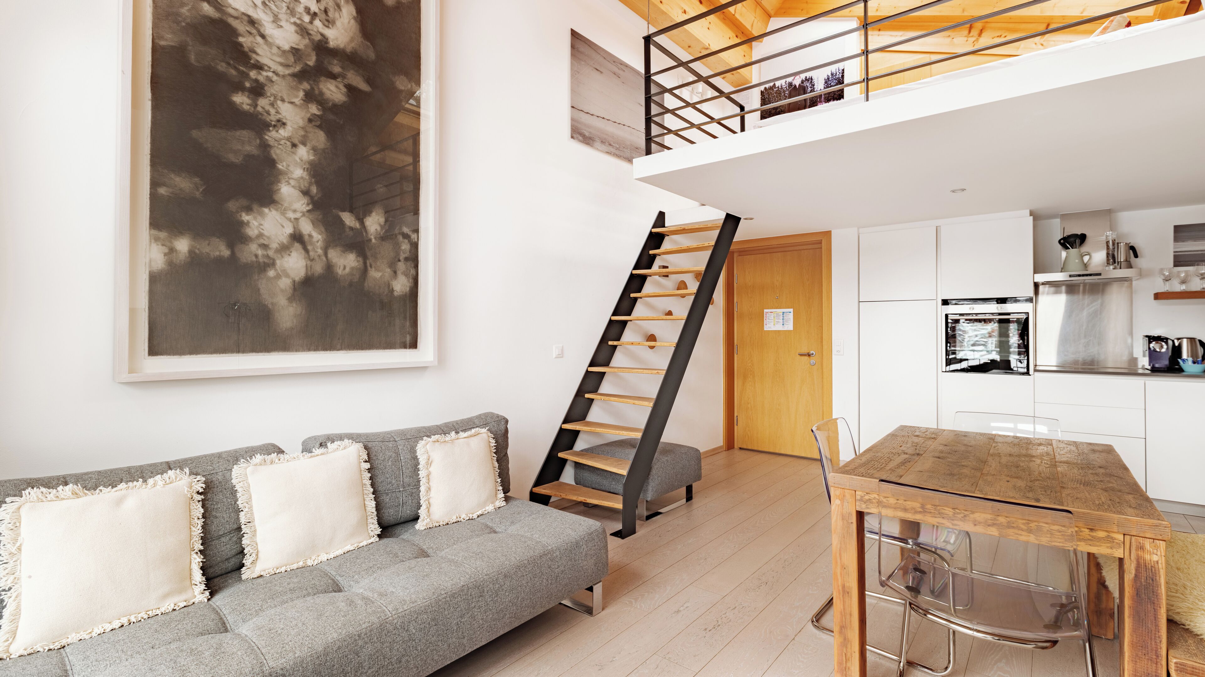 Een modern appartement met een open keuken in loftstijl, een grijze bank met witte kussens, een houten eettafel en een trap naar een hoger niveau.