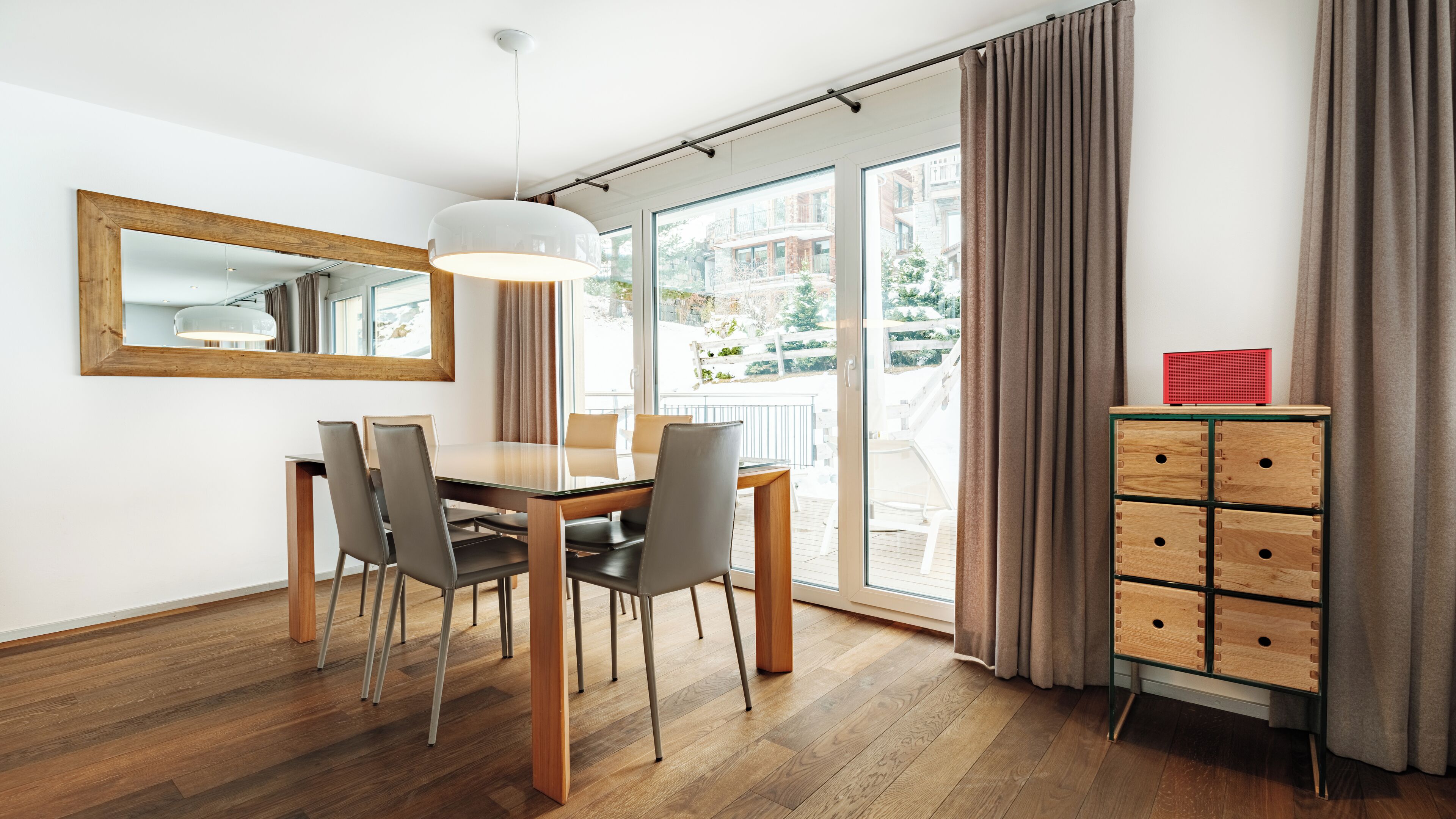 A modern dining area with a wooden table and chairs, a pendant light, a mirror, and a view of the outdoors.