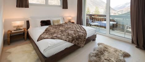 Un dormitorio moderno con una cama grande, un ventanal de vidrio que da a un balcón y vistas a montañas.