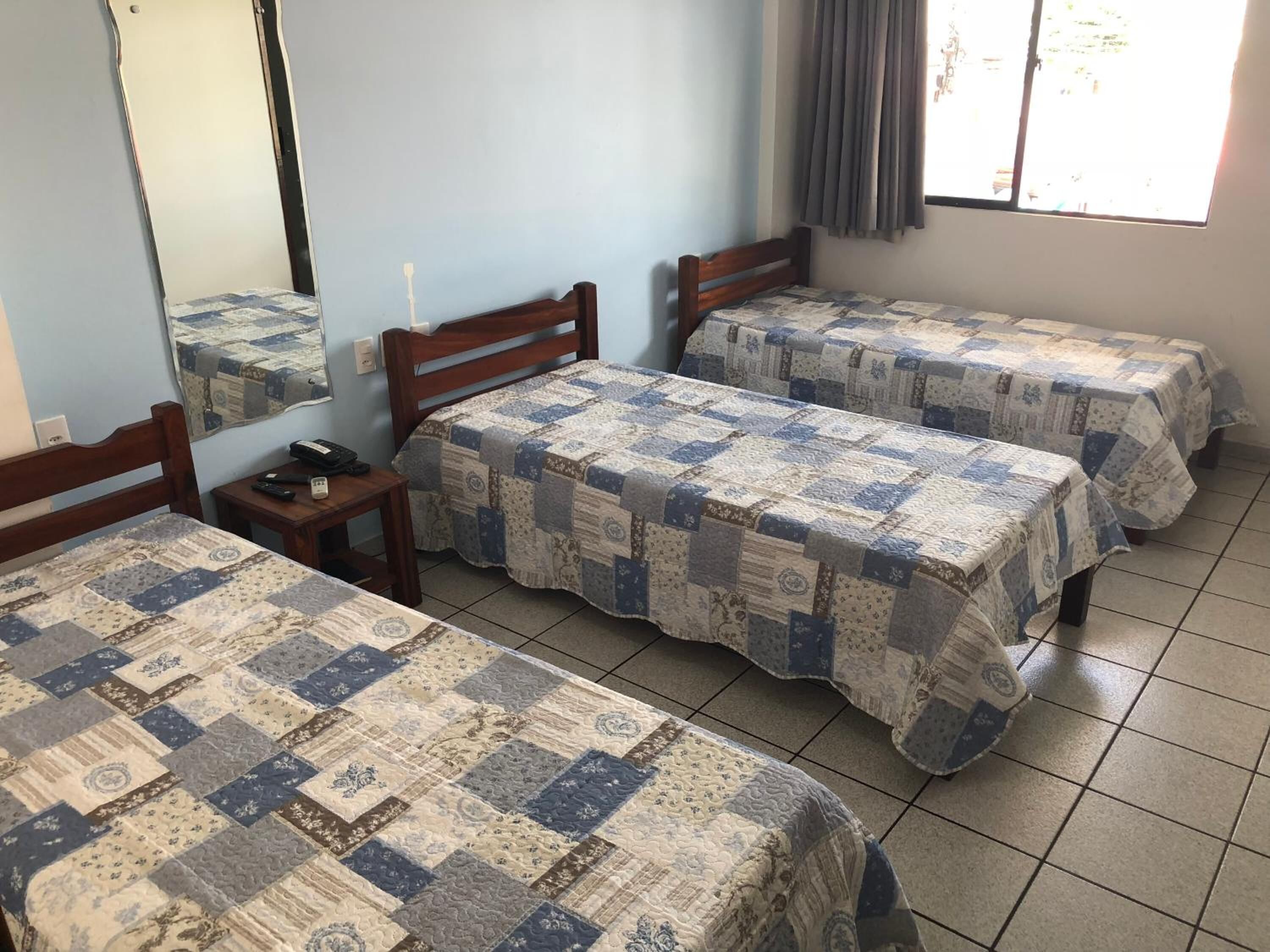 A room with three beds, each covered with a quilted bedspread, a wooden headboard, and a small bedside table with a phone and remote control.