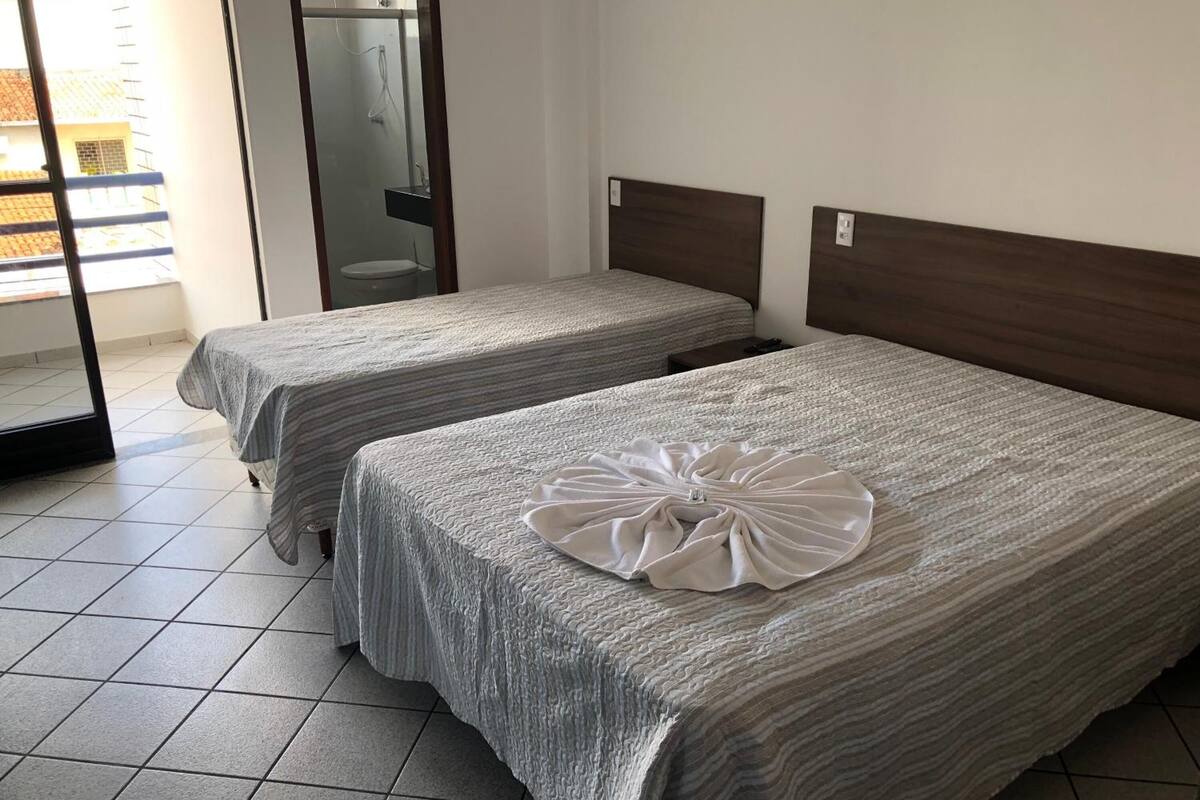 A hotel room with two beds, a bathroom with a glass door, and a view of a building outside.