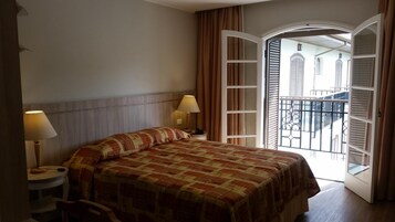 A hotel room with a bed, two bedside lamps, a small table, and a balcony with a view of buildings.