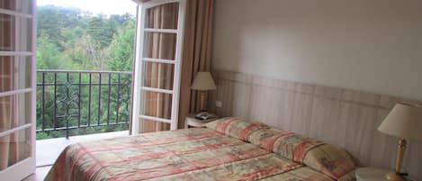 Un dormitorio con cama, dos mesitas de noche, una lámpara de noche, una ventana con cortinas y vistas a la vegetación.