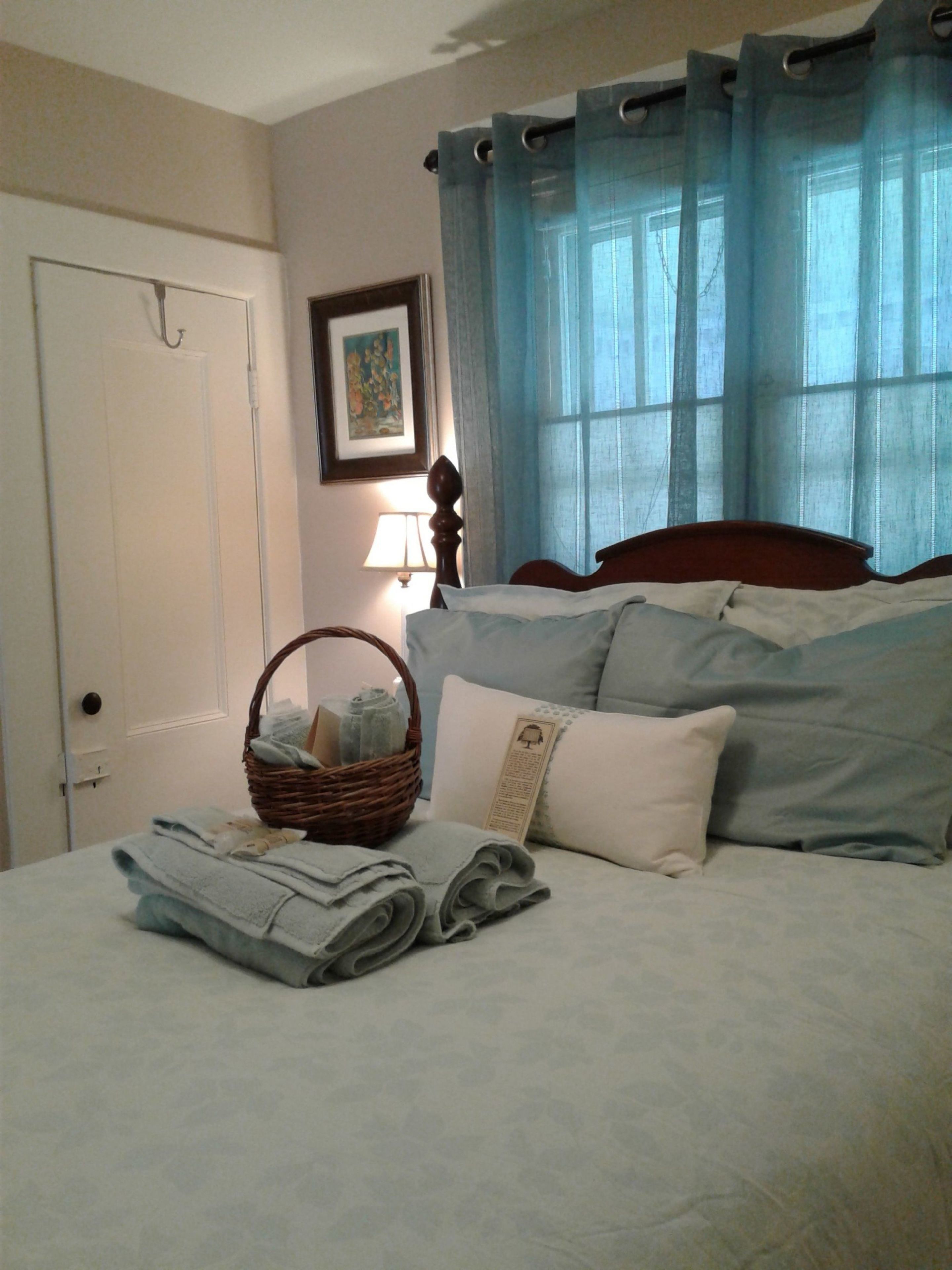 Una cama bien hecha con almohadas y toallas dobladas, una cesta de mimbre con ropa de cama, una lámpara de noche y una ventana con cortinas azules.