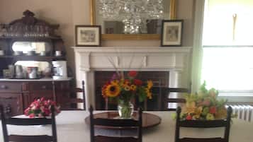 A dining room with a large table surrounded by four chairs, a chandelier above, and a fireplace with framed pictures above it.