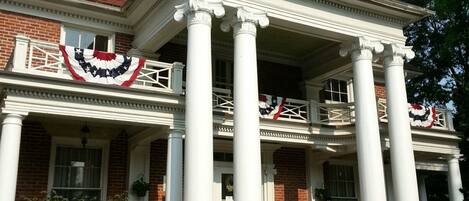 A two-story building with a central porch supported by columns, a brick facade, and a balcony with railings.
