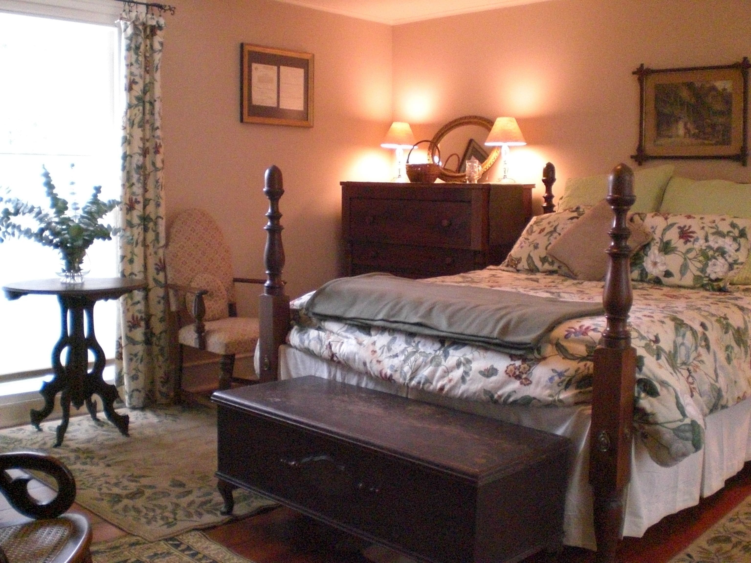 A bedroom with a four-poster bed, a nightstand, a chair, and a small table with a vase of flowers.