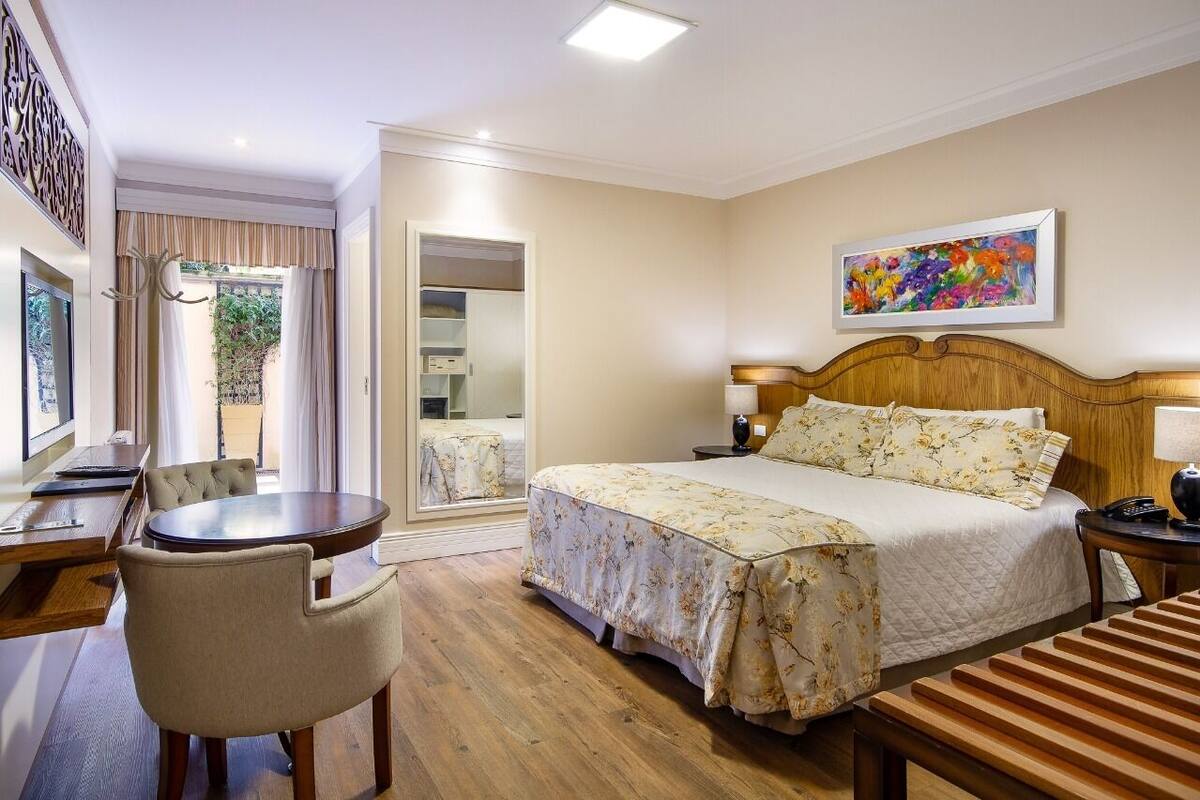 Una habitación de hotel con cama, mesita de noche, silla y escritorio con otra silla. Hay una ventana con cortinas, un cuadro en la pared y una puerta que da a otra habitación.