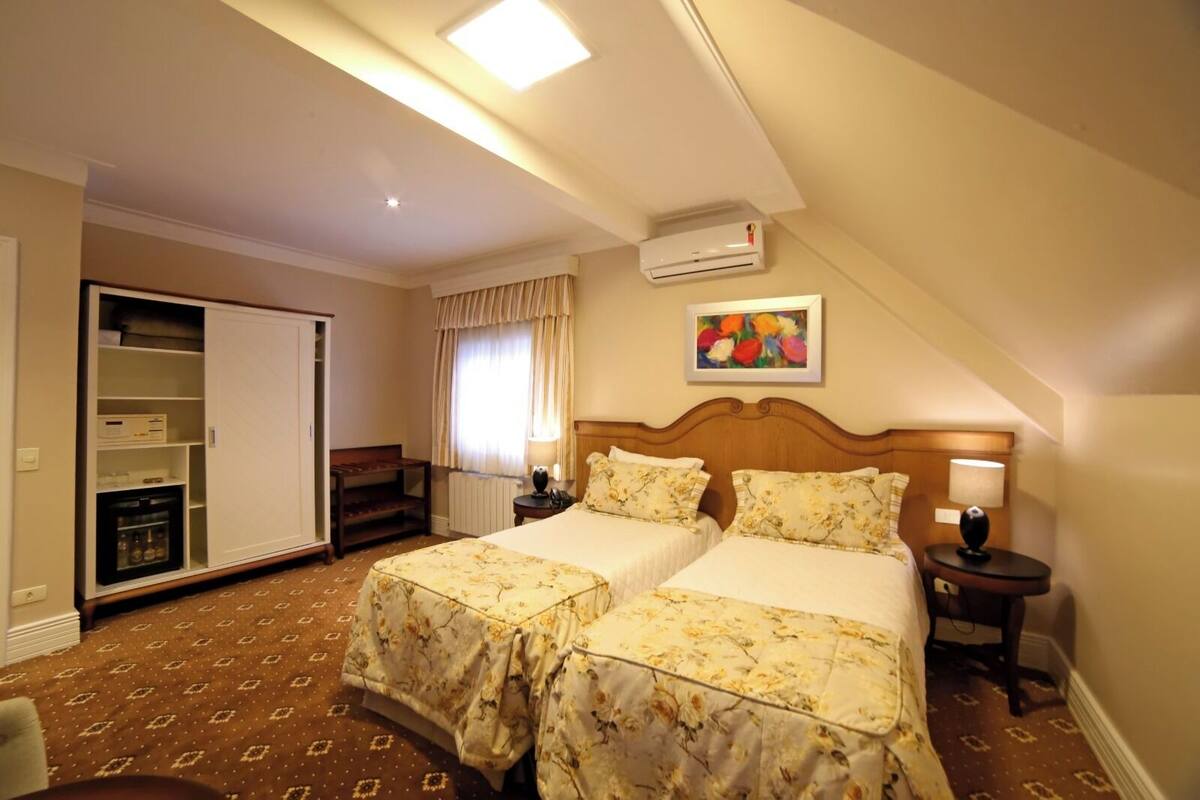 Habitación de hotel con dos camas, un armario, un escritorio, una silla, un equipo de aire acondicionado y un cuadro en la pared.