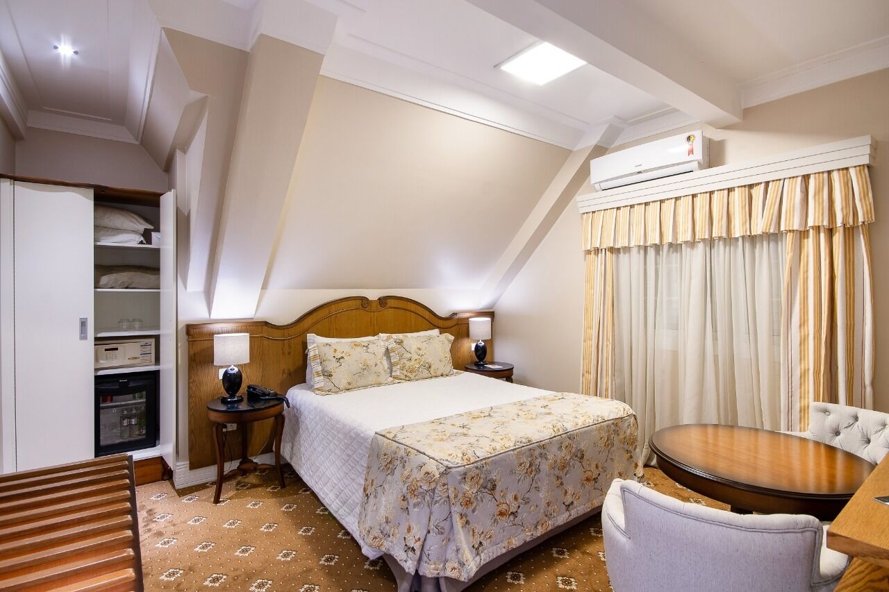 A hotel room with a bed, bedside tables, a small round table, a chair, a wardrobe, and a built-in air conditioning unit.