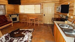 L’intérieur d’un chalet confortable en rondins, avec une cheminée, des meubles en bois, un coin cuisine et un ventilateur de plafond.