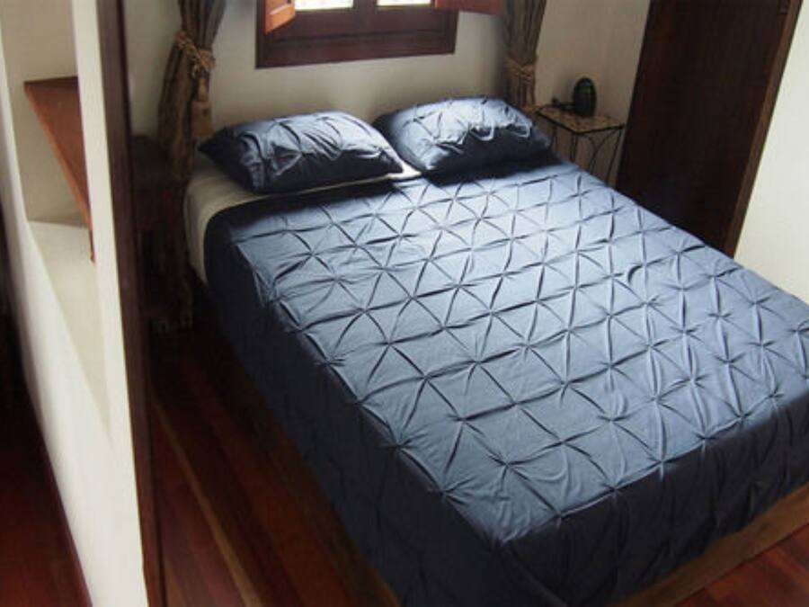 Een bed met een deken met een geometrisch patroon, een houten vloer, een raam met gordijnen en een kleine tafel met een lamp.