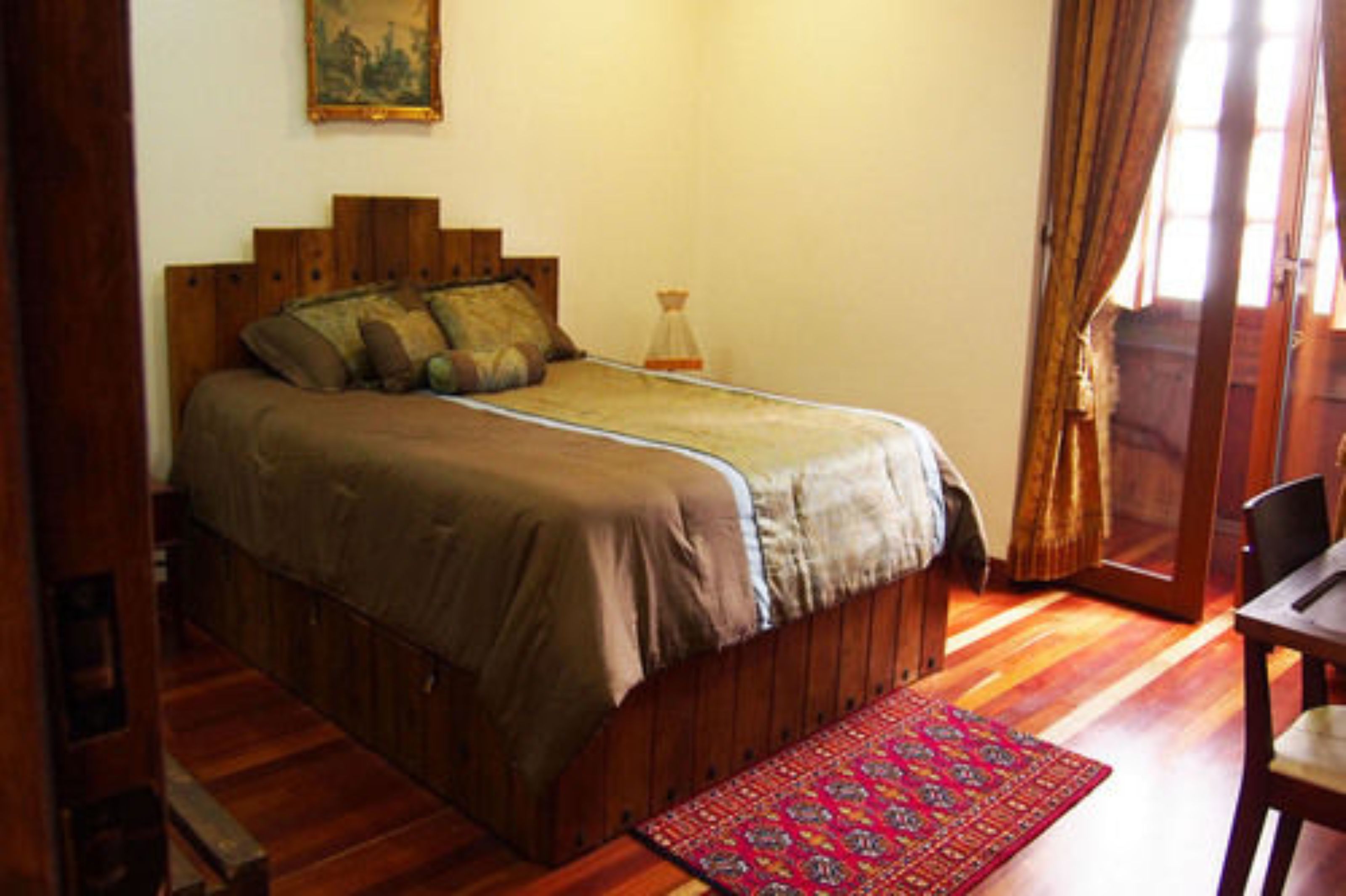 A bedroom with a wooden bed, a nightstand, a chair, a window with curtains, and a painting on the wall.
