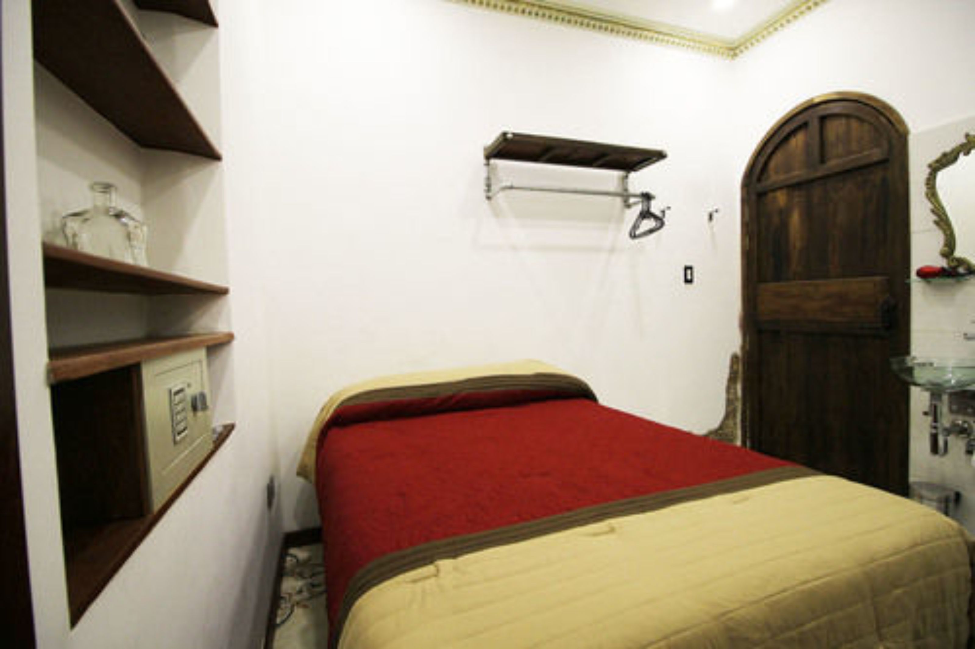 A small, narrow room with a bed, a wooden door, a shelf with a bottle, and a mirror on the wall.