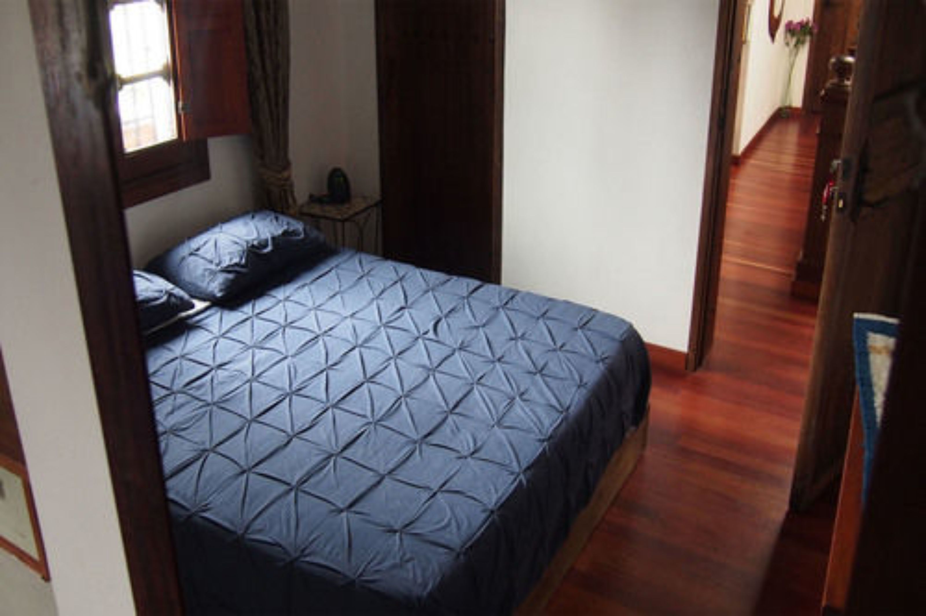 A small, narrow room with a single bed, a wooden floor, and a window with curtains.