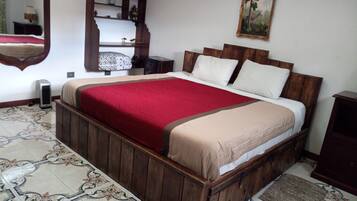 Une chambre à coucher avec un lit en bois, une couvre-lit rouge et beige, une table de chevet en bois, un miroir, une étagère avec des objets décoratifs et un sol à motifs.