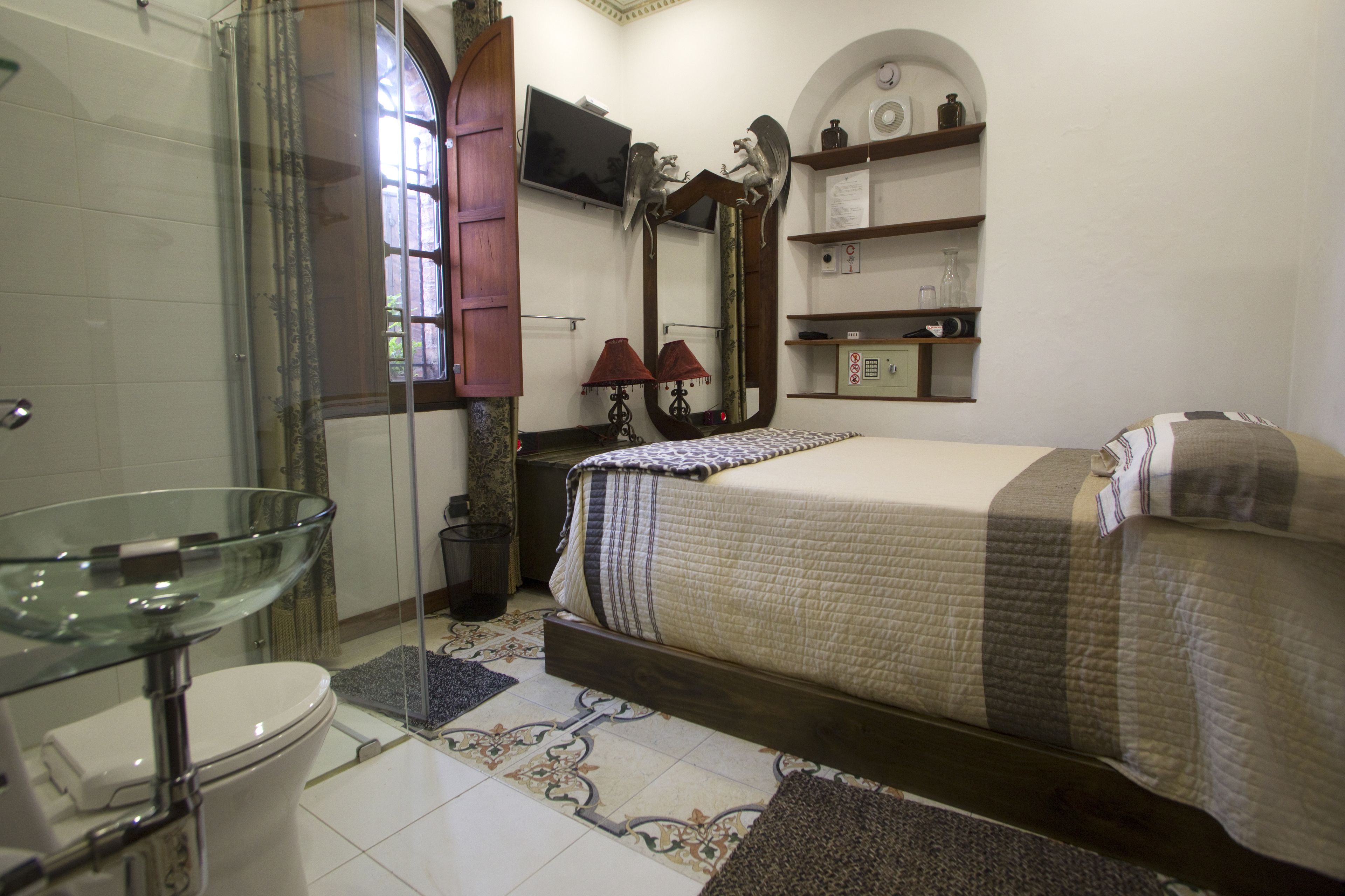 A bathroom with a glass shower enclosure, a toilet, and a bed with a patterned bedspread.