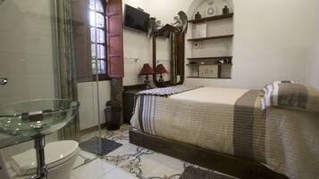 A bathroom with a glass shower enclosure, a toilet, and a bed with a patterned bedspread.