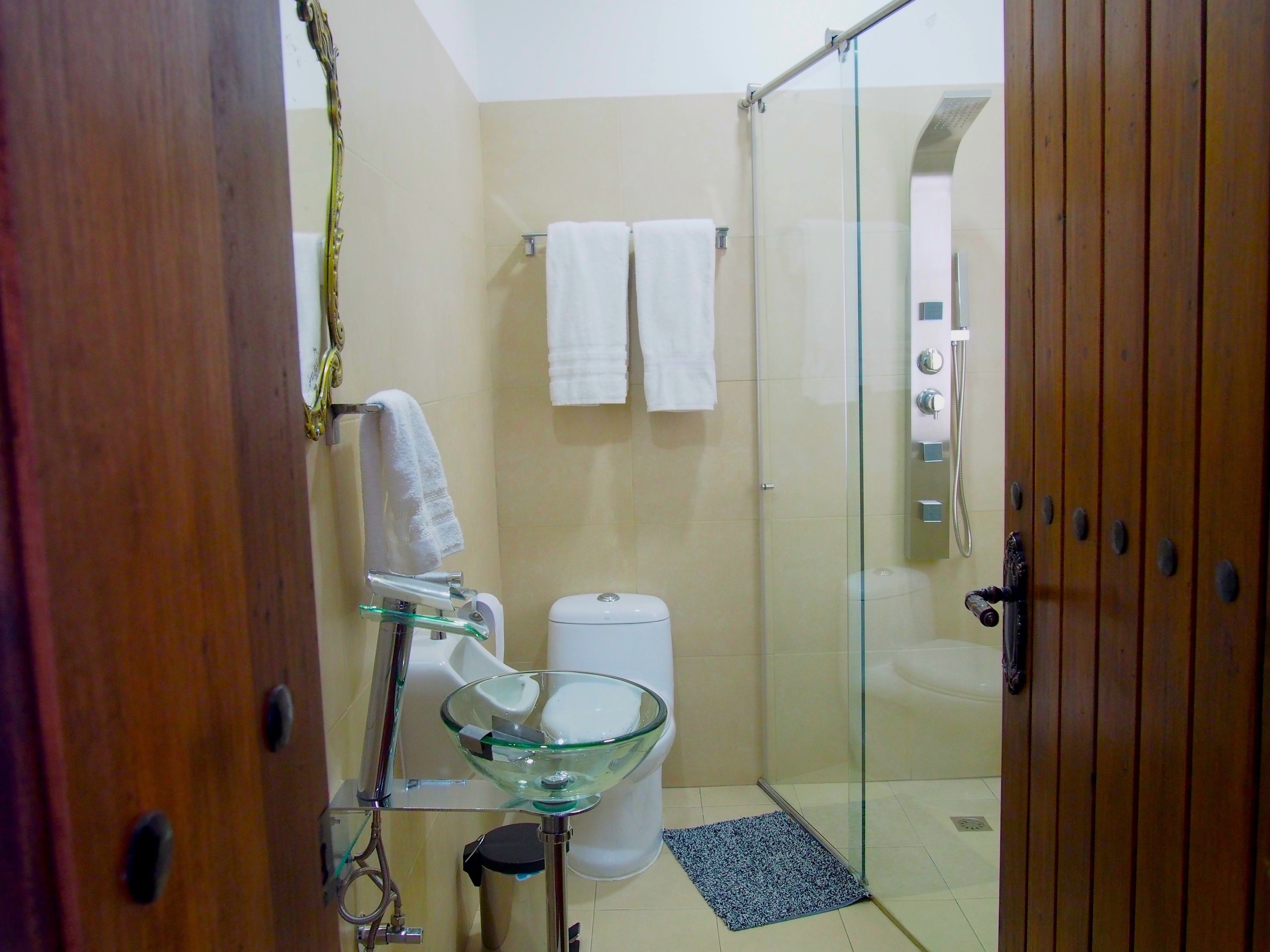 A bathroom with a glass shower enclosure, a glass sink, and a toilet. There are two white towels hanging on the wall.