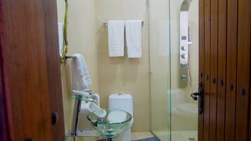 A bathroom with a glass shower enclosure, a glass sink, and a toilet. There are two white towels hanging on the wall.