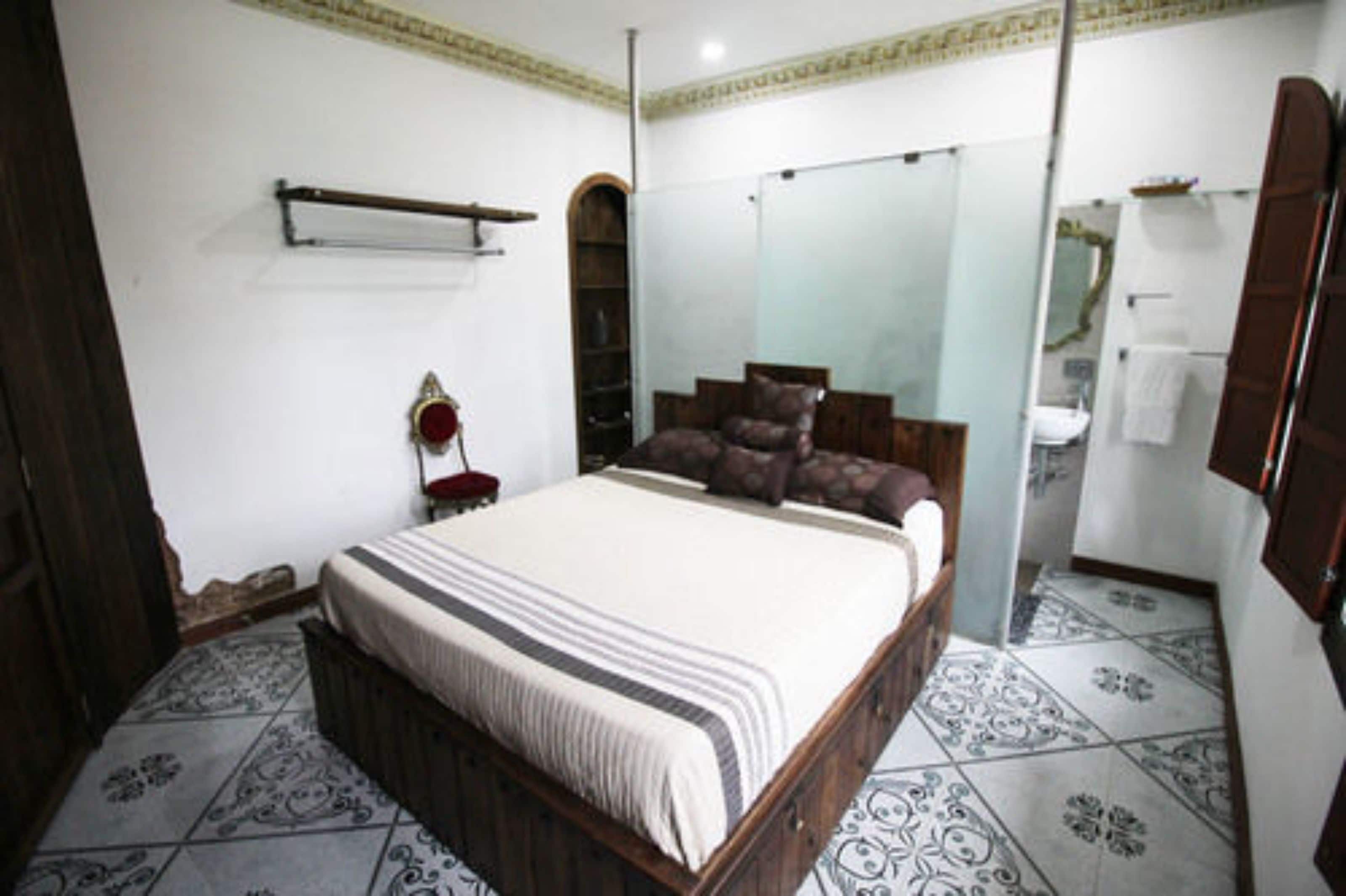 A bedroom with a large bed, a wardrobe, a mirror, and a bathroom visible through an arched doorway.