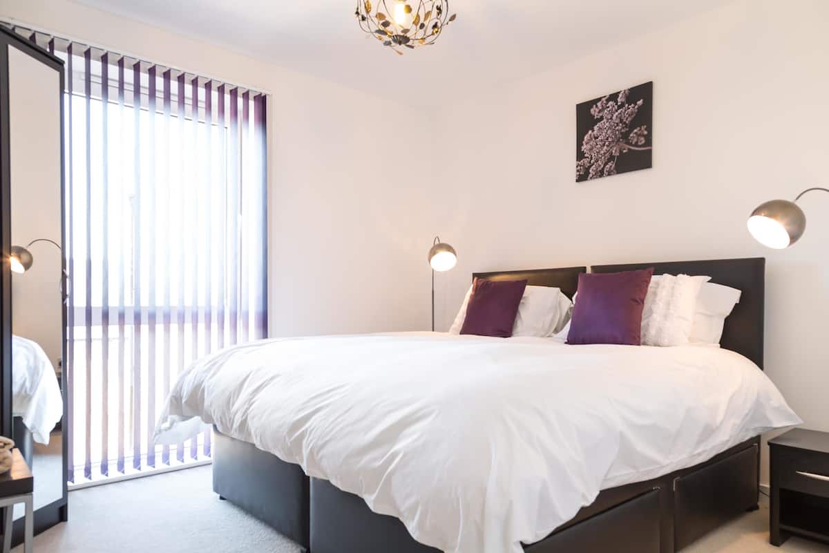 Une chambre moderne avec un grand lit, deux tables de chevet, un luminaire au plafond et une fenêtre avec des stores.