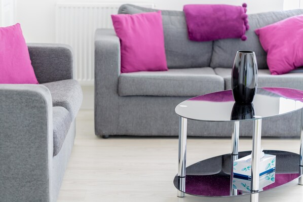 A modern living room with grey sofas and purple cushions, a glass coffee table with a black vase, and a tissue box on the table.