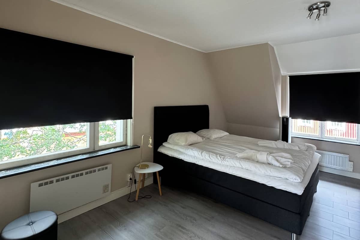 Un dormitorio con una cama, un taburete pequeño, un radiador y una ventana con persianas.