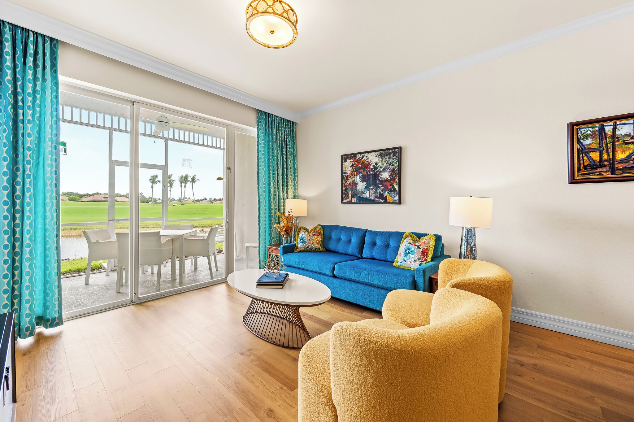 A living room with a blue sofa, yellow armchairs, a round coffee table, and a view of a golf course through large glass doors.
