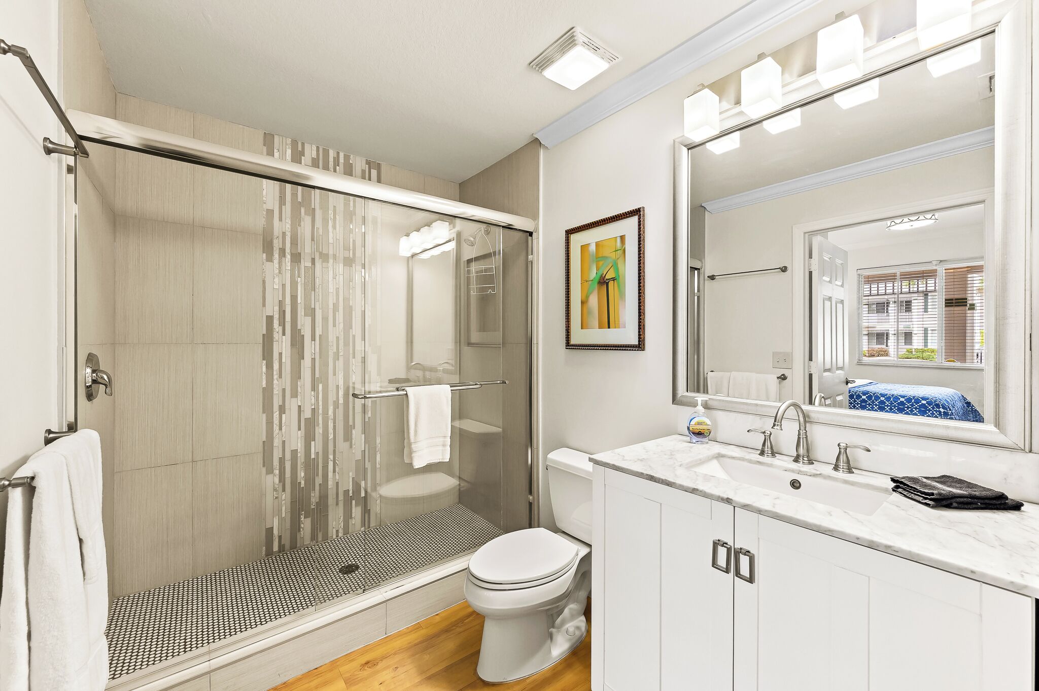 A modern bathroom with a glass shower enclosure, a white toilet, a sink with a mirror, and a framed artwork on the wall.