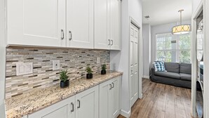 A kitchen with white cabinets, a granite countertop, and a mosaic tile backsplash. There is a gray sofa in the adjacent living area with a hanging light fixture. - Solterra Vacation Rental: Luxury 3-BR Condo with Lake & Golf Views. Access to Luxury Pool, Hot Tub, and Golf Courses at Lely Resort. Close to Naples Beaches, 5th Avenue S, and Marco Island (Naples)