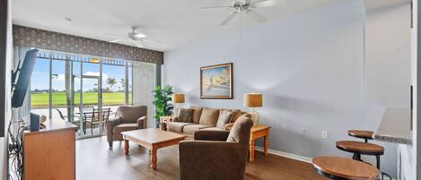 A spacious living room with a large window, a ceiling fan, a sofa set, a coffee table, and a small bar area.