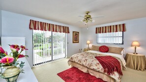 A bedroom with a bed, two bedside tables with lamps, a ceiling fan, a window with curtains, and a view of the outdoors.