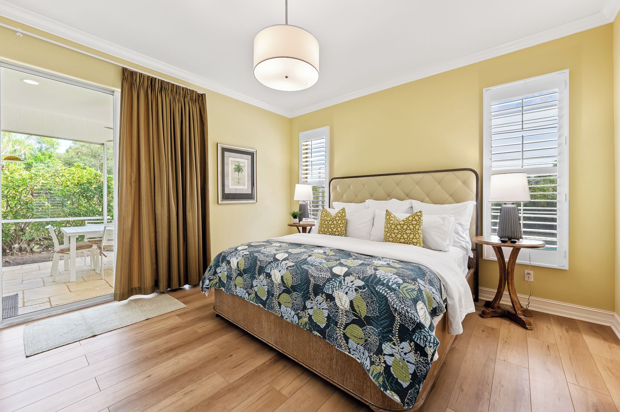 A bedroom with a bed, a bedside table, a lamp, a window with blinds, and a view of an outdoor patio.