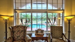 A room with two patterned armchairs, a small wooden table with a clock, and a large window with a view of a pool and palm trees.