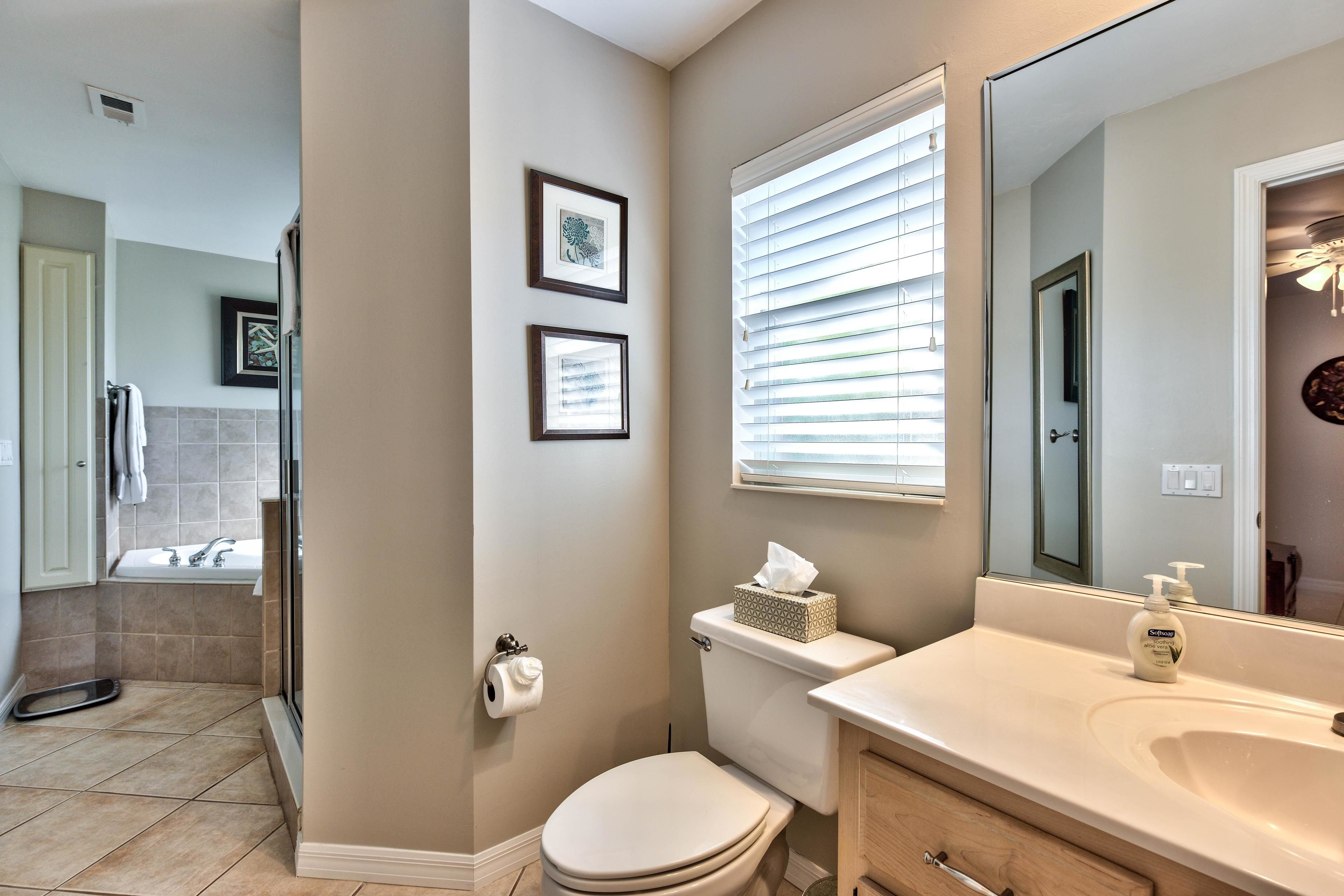 Une salle de bain avec des toilettes, un lavabo et un grand miroir. Deux tableaux encadrés ornent le mur, et il y a une fenêtre avec des stores.