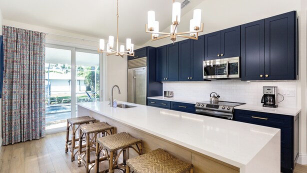 A modern kitchen with dark blue cabinets, a white countertop, and a stainless steel range hood. There are three wicker bar stools and a large window with a view of the outdoors. - Tuscan Vacation Rental. Stylish 3-Bedroom Getaway Near Vanderbilt Beach Enjoy a Private Pool, Spacious Layout, and Modern Comforts Perfect for Families, Groups, and Pet-Friendly Stays (Naples)