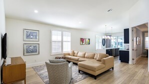 A spacious living room with a beige sectional sofa, a patterned ottoman, and a wooden coffee table. There are framed artworks on the walls, a large window with blinds, and a kitchen area visible through an open door. - Tuscan Vacation Rental. Stylish 3-Bedroom Getaway Near Vanderbilt Beach Enjoy a Private Pool, Spacious Layout, and Modern Comforts Perfect for Families, Groups, and Pet-Friendly Stays (Naples)