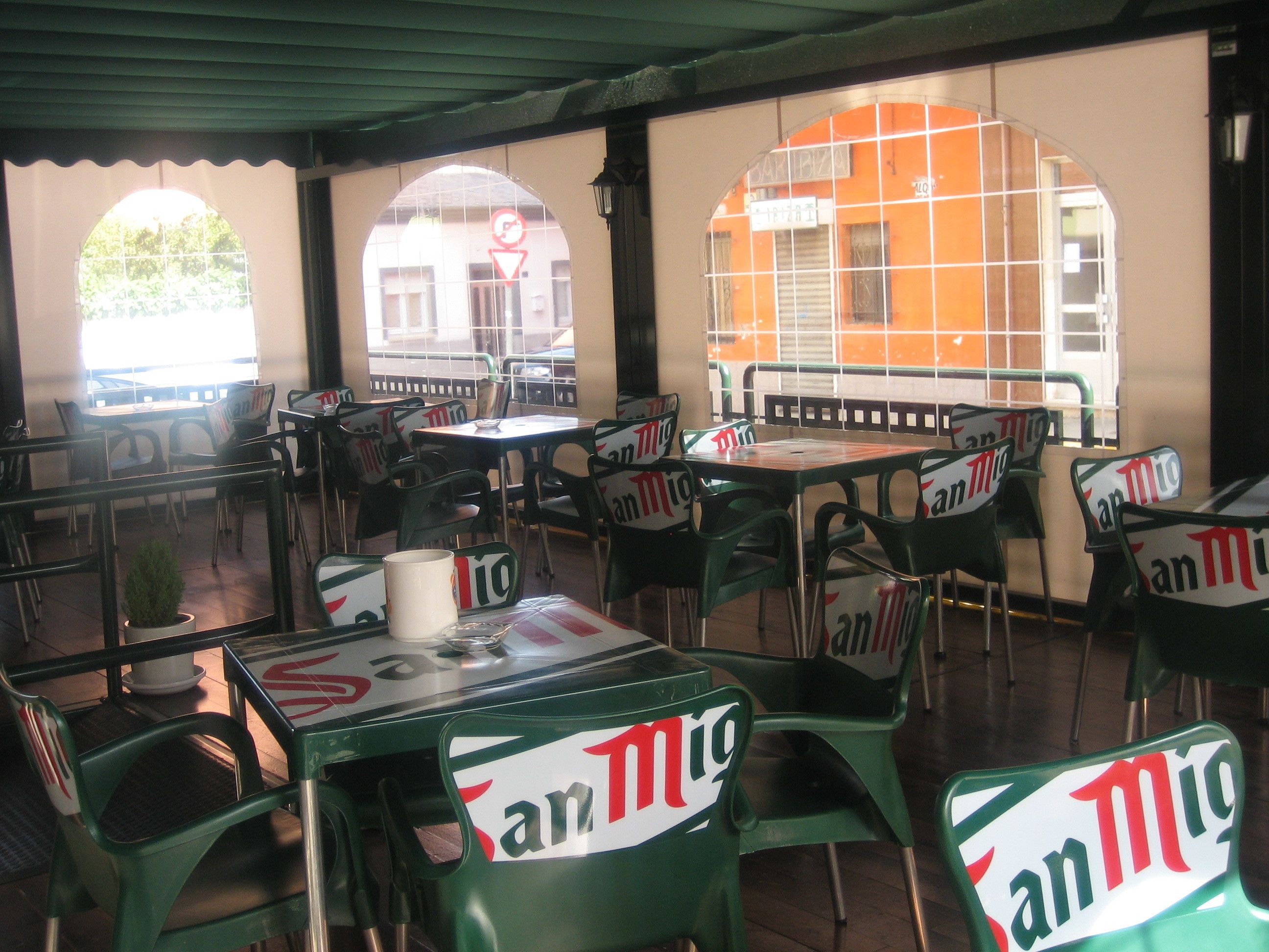 Un café al aire libre con mesas y sillas, algunas con la marca 'San Mig'.