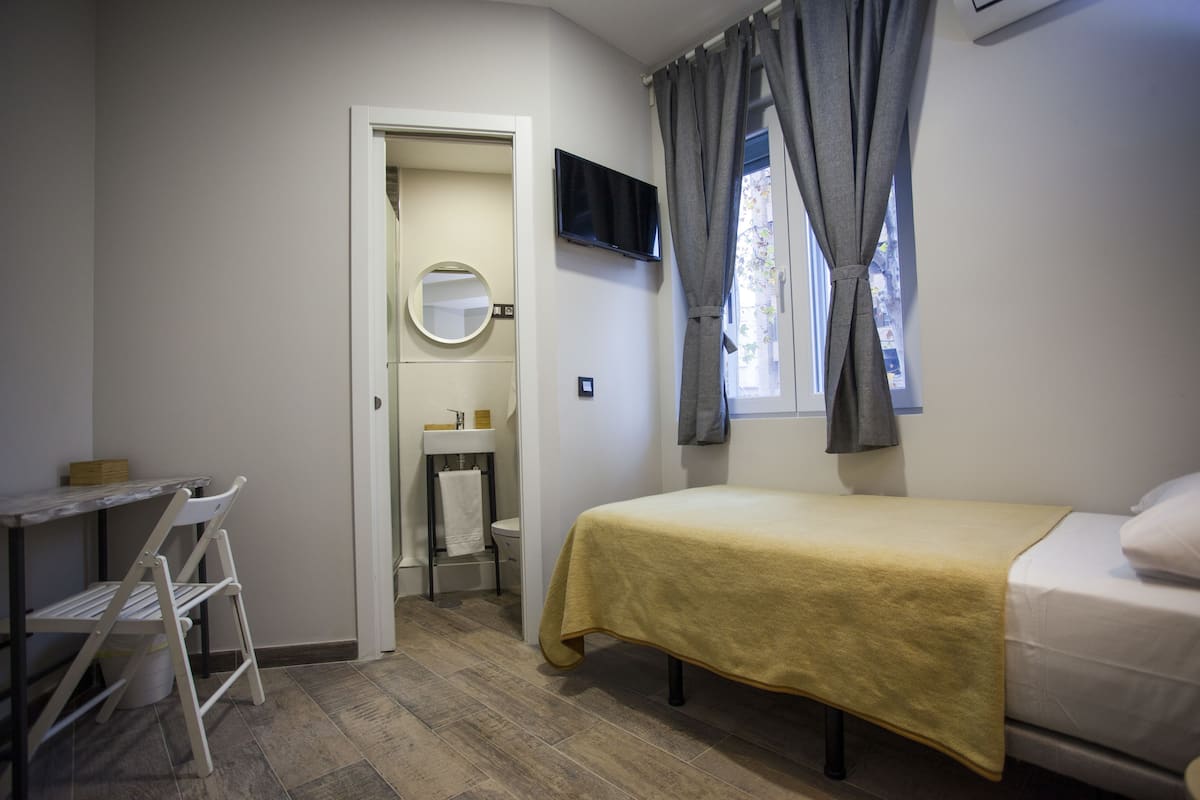 A bedroom with a bed, a desk, a chair, a mirror, a TV, and a window with curtains.