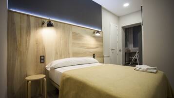 Un dormitorio con cama, mesita de noche y un taburete pequeño. Se aprecia una ventana con cortinas y una puerta que da a otra habitación.
