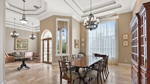 A spacious dining room with a large wooden table and chairs, elegant chandeliers, and a view of the outdoors through large windows. - Knickerbocker Estate: Elegant 9-BR Getaway in Naples. Enjoy Private Pool, Outdoor Dining, and Expansive Grounds, Experience Tranquility with Easy Access to Naples' Best (Naples)