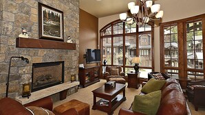 Un salon chaleureux avec une cheminée en pierre, des meubles en cuir et de grandes fenêtres donnant sur un chalet en bois.