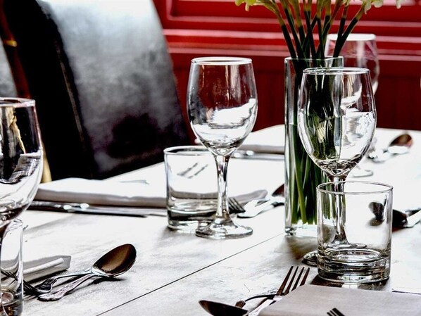 Una mesa de comedor con cristalería transparente, incluyendo copas de vino y vasos para agua, y un jarrón alto con tallos verdes.
