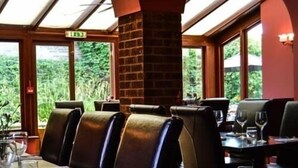 A restaurant with leather booths, a brick pillar, and large windows offering a view of greenery. - The Crown Hotel (Sudbury)