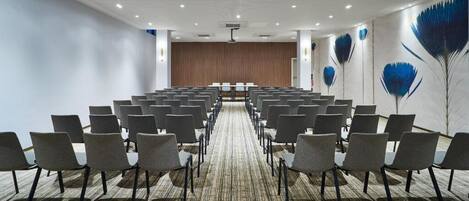 Un salón de conferencias con hileras de sillas, piso alfombrado y un escenario al frente.