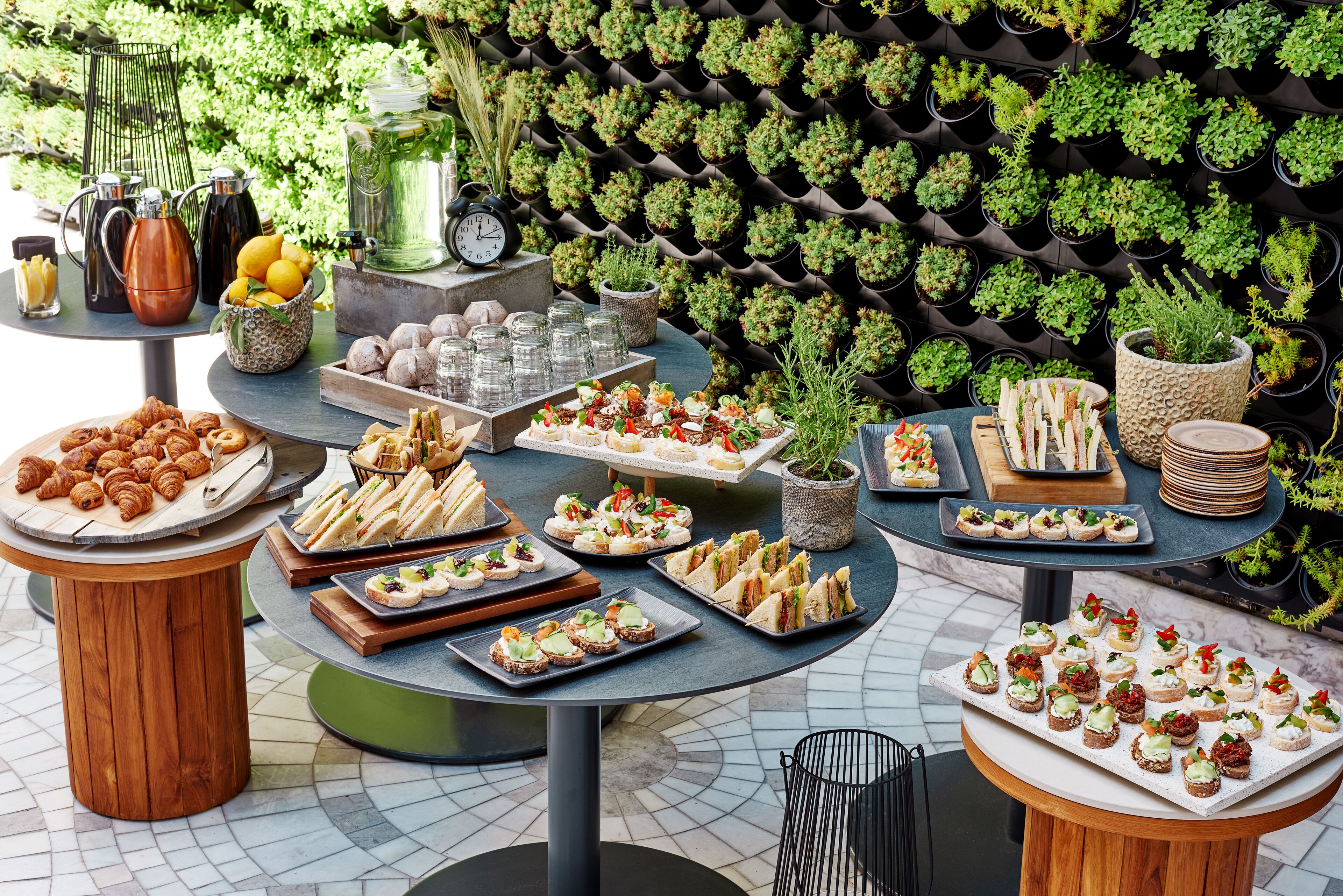 A variety of food and drink items displayed on tables outdoors, with a backdrop of a wall-mounted plant arrangement.