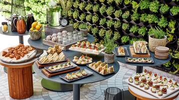 A variety of food and drink items displayed on tables outdoors, with a backdrop of a wall-mounted plant arrangement.
